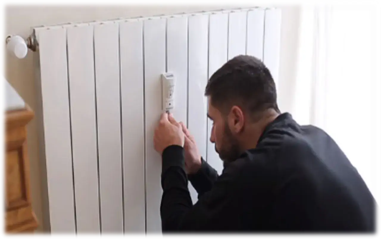Hombre ajustando un dispositivo en un radiador blanco. Estos son contadores individuales para calefacci&oacute;n central, permitiendo un control eficiente.