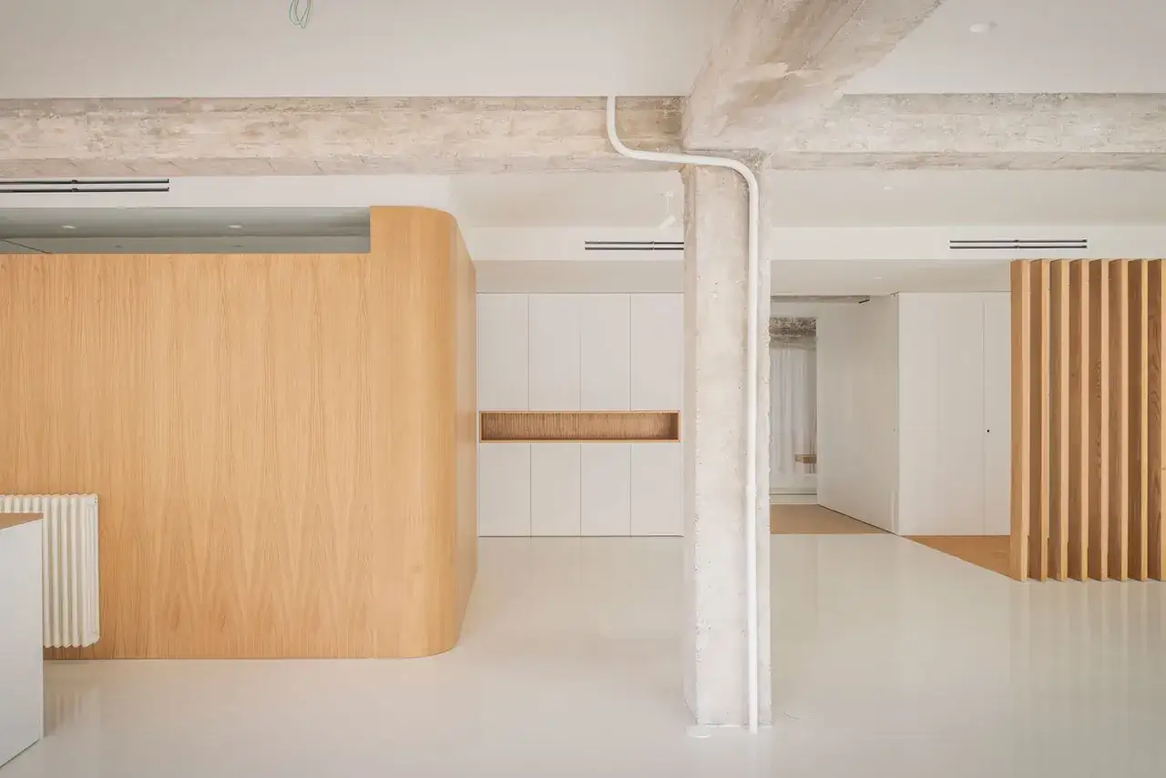 Espacio moderno con paredes curvas de madera y armarios blancos. El suelo es de resina y se aprecian vigas de hormig&oacute;n, creando un ambiente con revestimientos continuos.