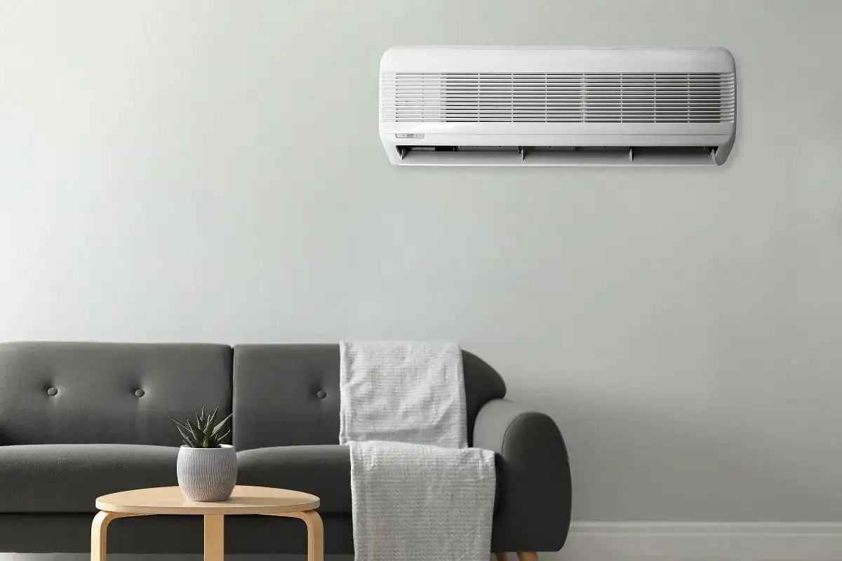 Unidad de aire acondicionado montada en la pared, cumpliendo la normativa aire acondicionado. Sofá gris con mantas y mesa auxiliar con planta.