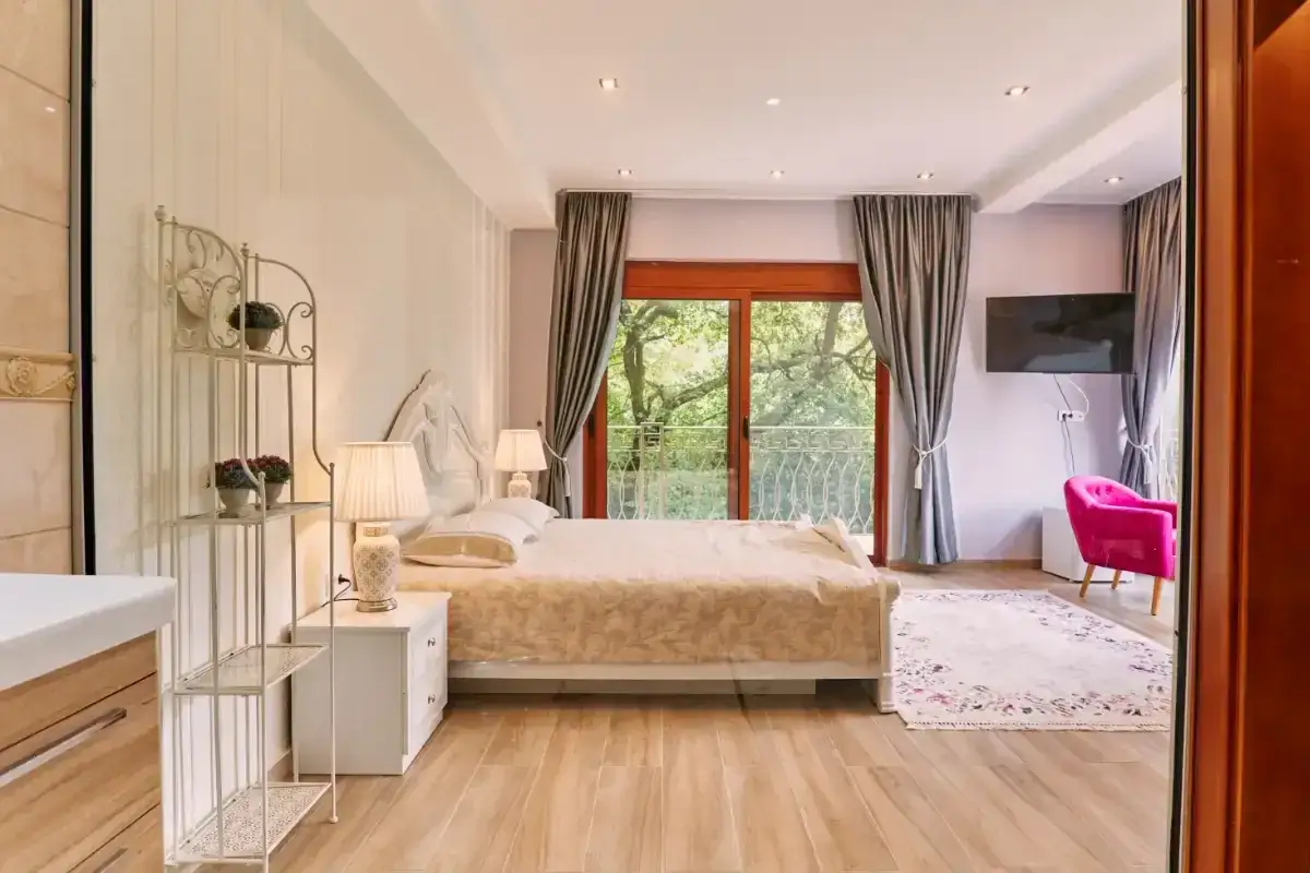 Habitaci&oacute;n con cama matrimonial, balc&oacute;n con vista a &aacute;rboles y un sill&oacute;n rosa. Se planea tirar tabique para unir dos habitaciones.