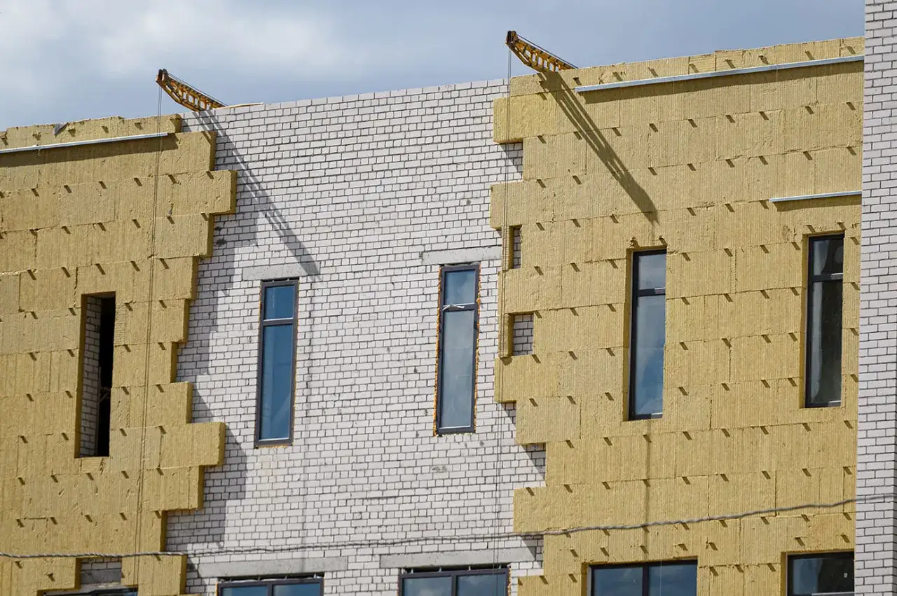 Fachada de edificio en construcci&oacute;n, con aislamiento de lana mineral amarilla y ladrillo blanco. Se aprecian ventanas y una gr&uacute;a en la parte superior.