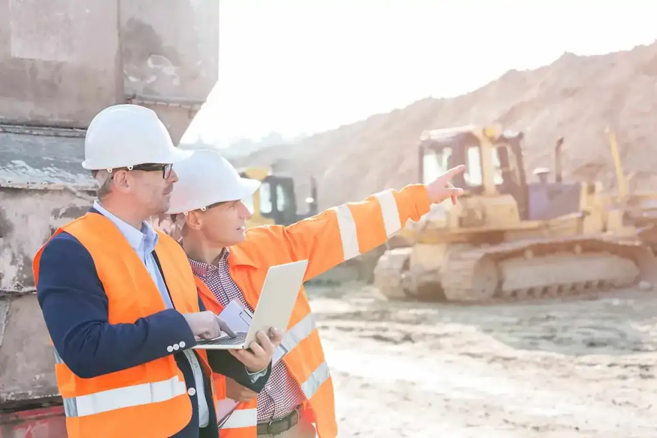 Dos ingenieros con cascos blancos y chalecos naranjas revisan un portátil. Uno señala a lo lejos, indicando el progreso de la obra.