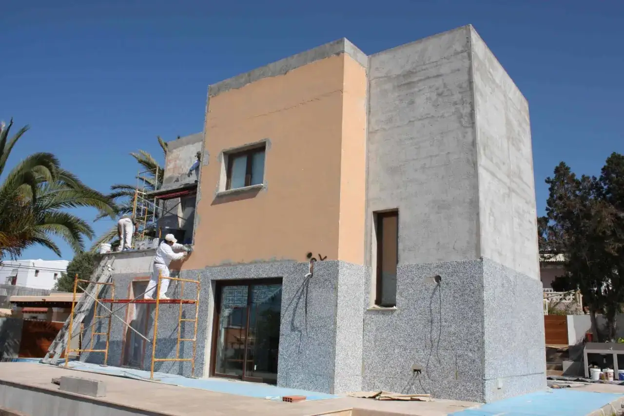 Obreros aplican un acabado sate a la fachada de una casa en construcción, con andamios y palmeras alrededor.