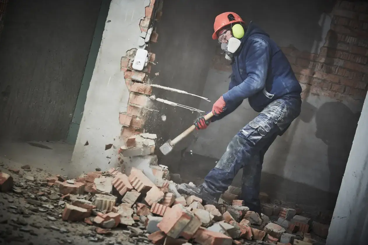 Obrero con casco y mascarilla demole un tabique de ladrillo con un mazo. Es un ejemplo de como tirar tabiques de ladrillo de forma segura.