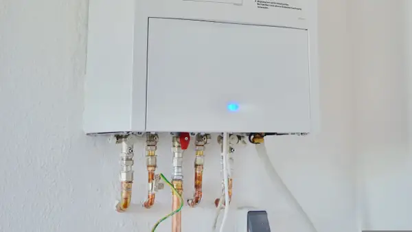 Calentador atmosférico blanco montado en pared con tuberías de cobre y conexiones. Una luz azul parpadea en la unidad, indicando que se puede instalar un calentador atmosférico en el interior.